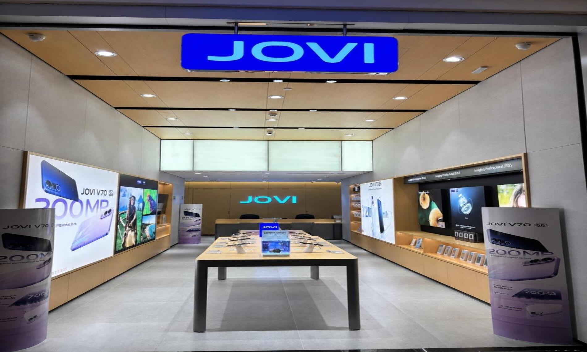 Loja conceito da JOVI com iluminação moderna, mesas centrais exibindo smartphones e grandes painéis promocionais do modelo JOVI V70 nas laterais.